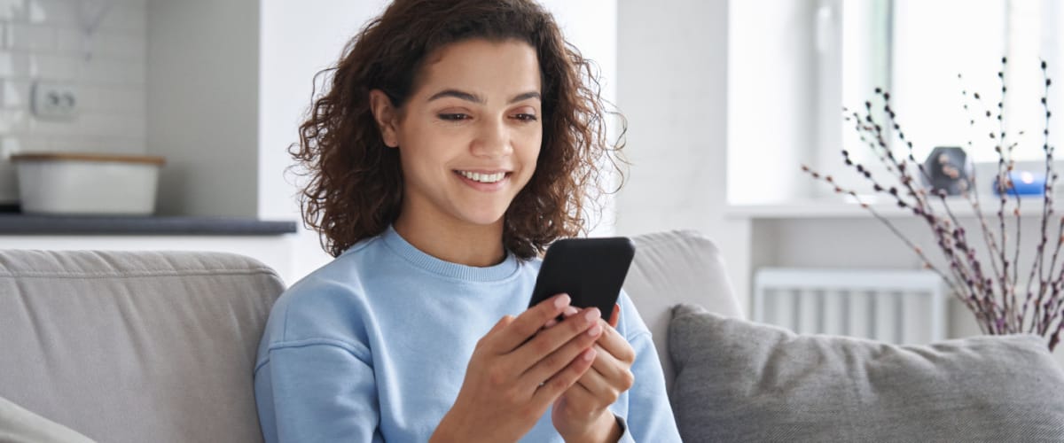 Happy hispanic teen girl holding cell phone using smartphone device at home. Smiling young latin woman blogger subscribing new social media, buying in internet, ordering products online using Dosh app